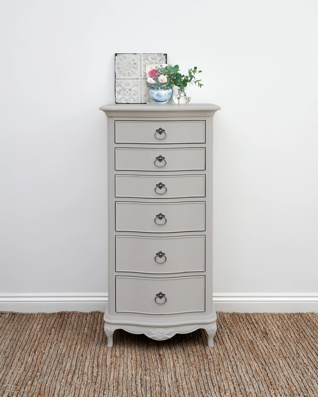 A tall, narrow gray painted chest of drawers with six drawers and ornate ring pulls, styled on a woven jute rug against a white wall.