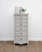 A tall, narrow gray painted chest of drawers with six drawers and ornate ring pulls, styled on a woven jute rug against a white wall.